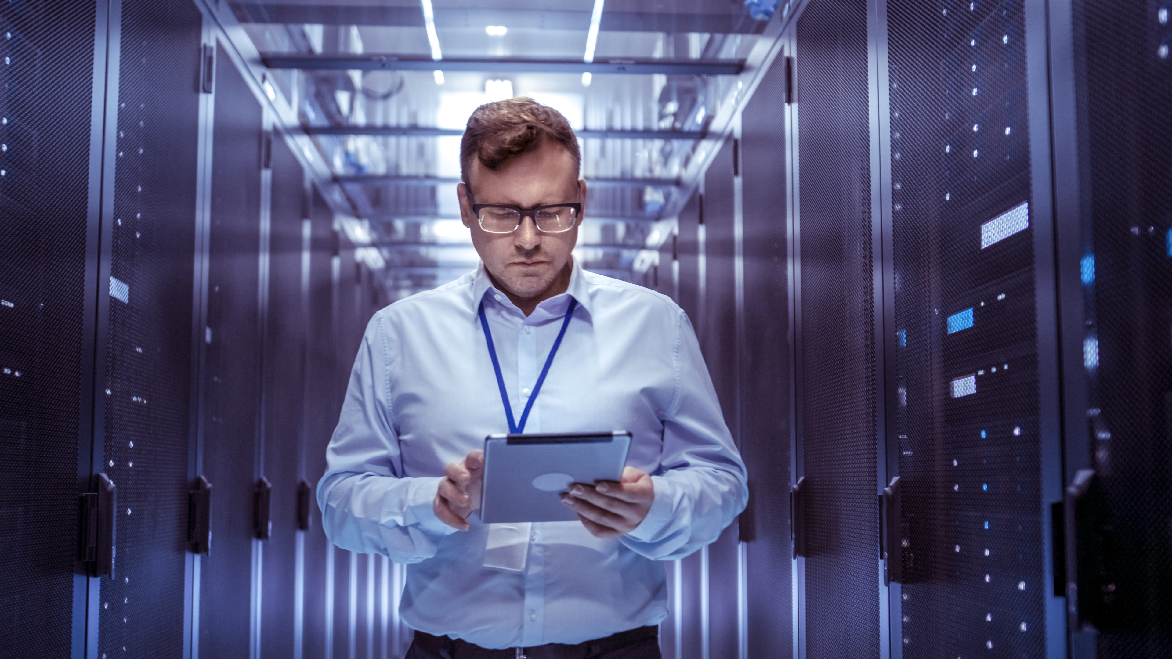 IT Technician Walks Through Rows of Server Racks in Data Center. Simultaneously He Works on a Tablet Computer.