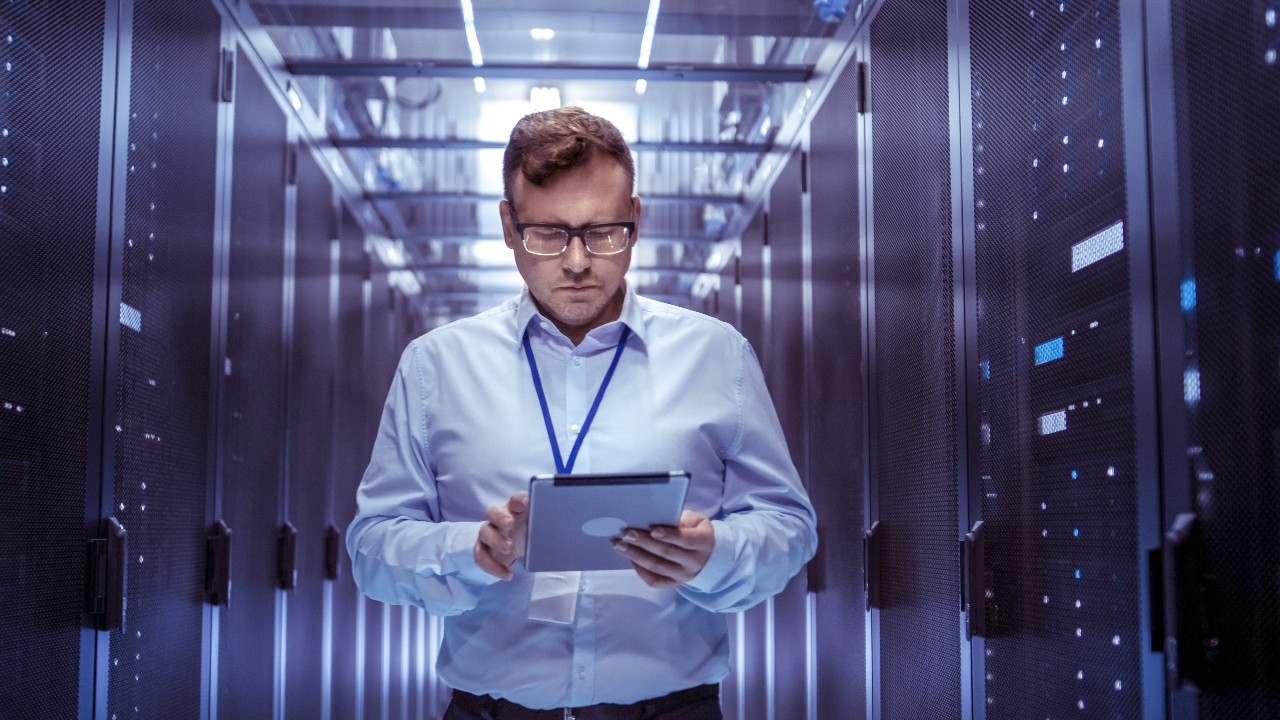 IT Technician Walks Through Rows of Server Racks in Data Center. Simultaneously He Works on a Tablet Computer.